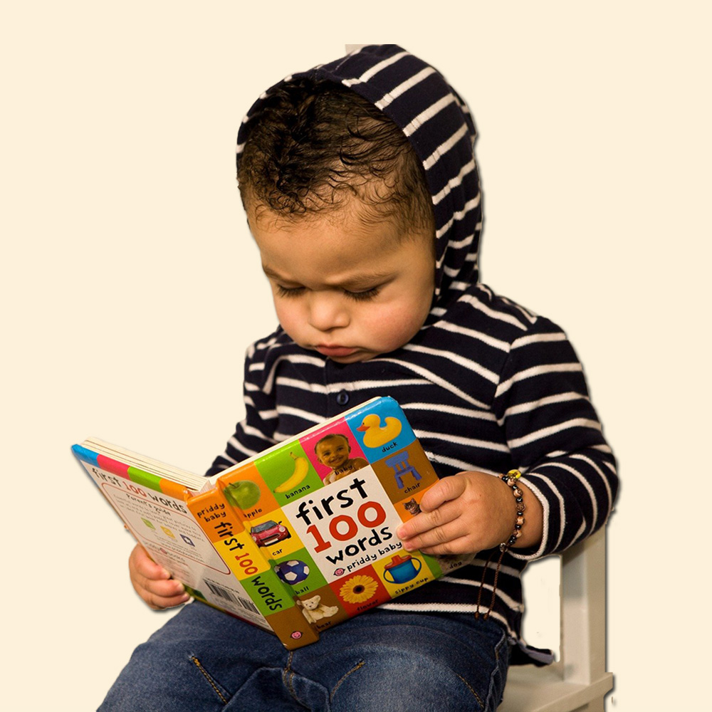 toddler wearing striped hooded shirt sitting in a chair reading a book of first words